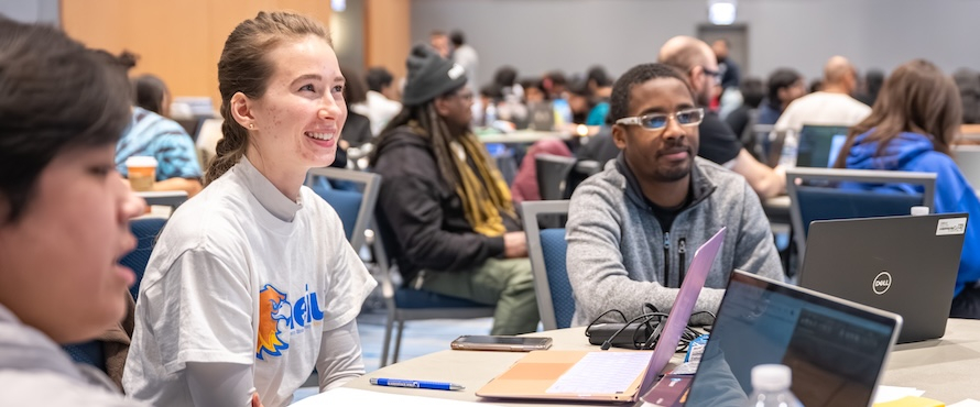 A photo of people sitting at tables with laptops open in Northeastern Illinois University's Alumni Hall. One is wearing a Northeastern Illinois University T-shirt.