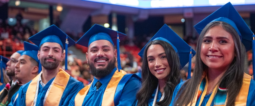A photo of students at at Northeastern Illinois University Commencement wearing blue caps and gowns with yellow stoles, smiling.