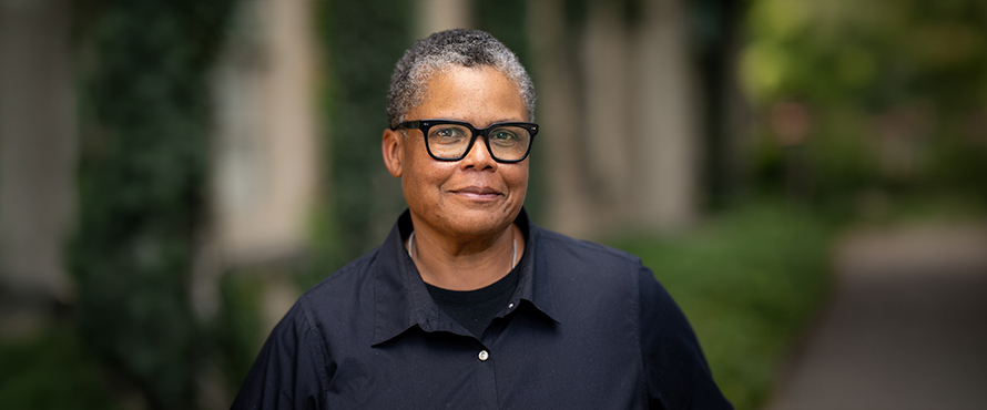 A headshot photo of Keeanga-Yamahtta Taylor, Ph.D., wearing black rimmed glasses and a dark blue collard shirt, smiling, with greenery in the background.