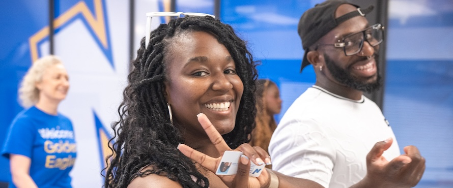 A photo of people walking down a hallway. The Northeastern Illinois University Flying N logo is in the background. Two people are smiling toward the camera. One is holding their NEIU ID card as they make a peace sign with their fingers.