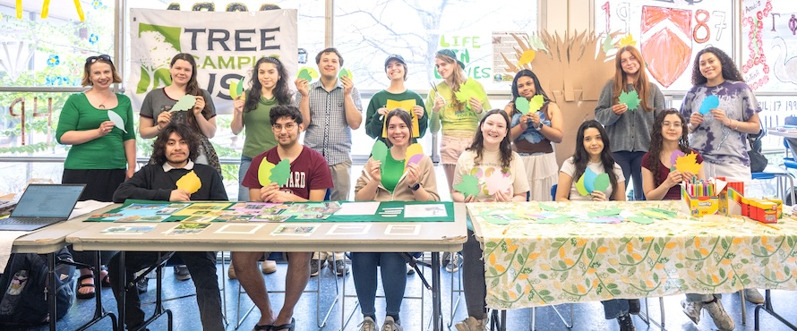 A photo of members of the Northeastern Illinois University community celebrating Arbor Day 2025 by hosting an informational table in Village Square on the Main Campus. A group of 15 people are holding paper leaves and smiling. 