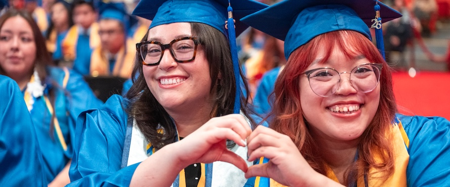 A photo of two Northeastern Illinois University students in blue graduation caps and gowns, smiling, making a heart shape with their hands.