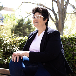 Dr. Davis is wearing a wihte eye glass, white shirt and navy suit jacket. She is seated and surrounded by trees.