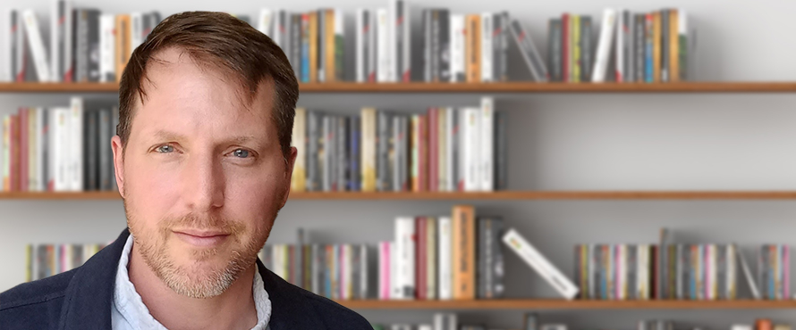 A headshot photo of NEIU Professor of History Joshua Salzmann, Ph.D., with shelves with books in the background.