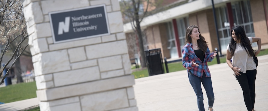 A photo of two females, smiling, in casual clothes and with backpacks, walking along the University Commons on a sunny day. A Northeastern Illinois University sign is visible, but blurred, in the foreground.