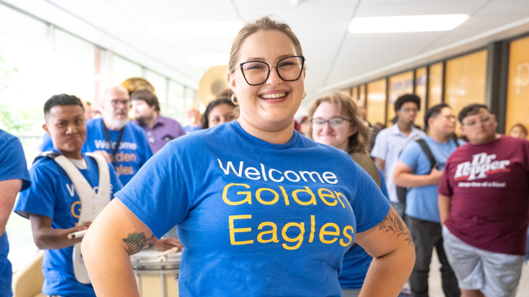 A photo of someone smiling wearing a blue shirt with text that reads "Welcome Golden Eagles" surrounded by people in the hallway of the E Building on Northeastern Illinois University's Main Campus.