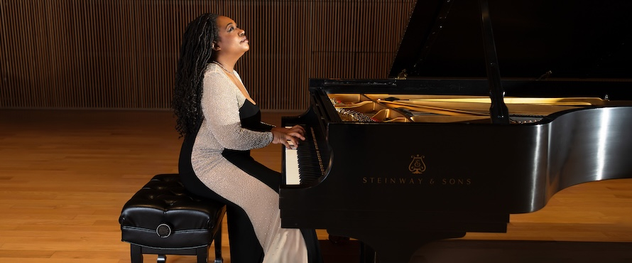 A photo of Michelle Cann sitting at a piano with her head tilted upwards. She is wearing a black and white dress.