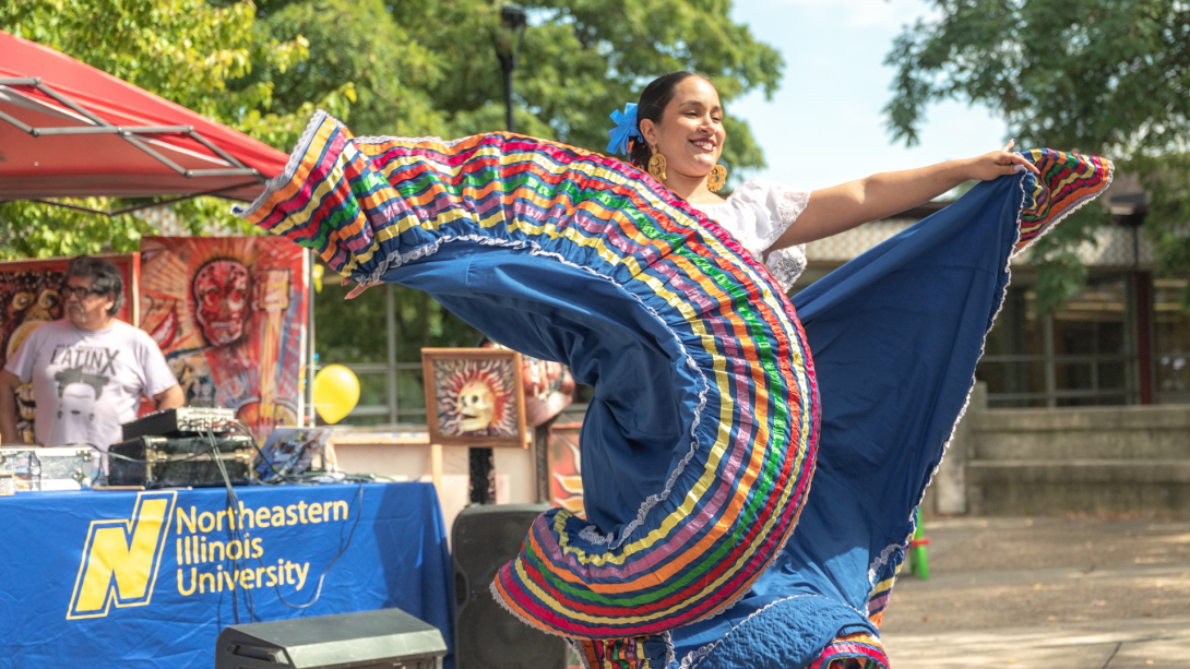 A photo of a folkloric dancer in a blue skirt with rainbow trim dancing at Northeastern Illinois University's Main Campus outside on a sunny day. 