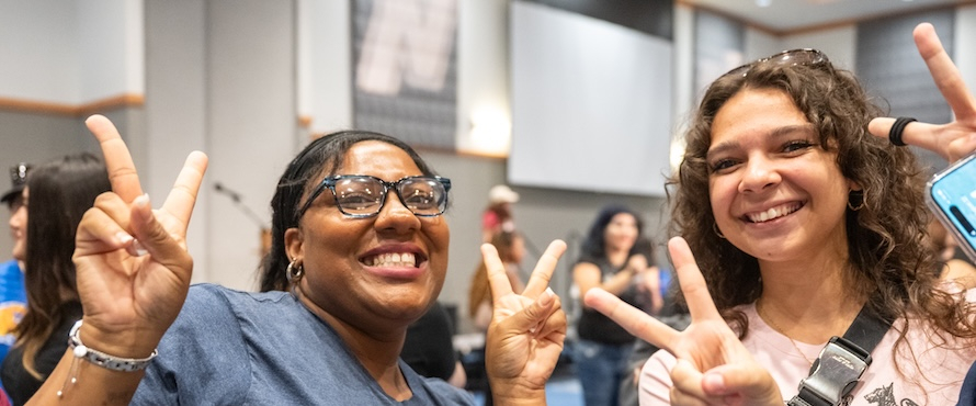 A photo of two people in Northeastern Illinois University's Alumni Hall. They are smiling and making peace signs with their hands.