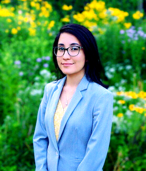 Obsmara Ulloa wearing a light blue suit jacket stands outdoors with flowers and greenery in the background