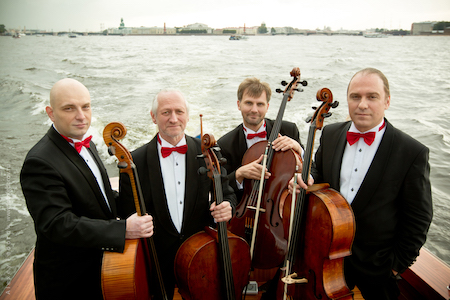 The quartet stands in a boat wearing tuxedos and holding their cellos.